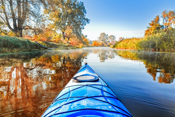 View from the blue kayak on the river banks with autumnal yellow leaves trees in fall season. The Seversky Donets river, autumn kayaking. View over nose of bright yellow kayak.